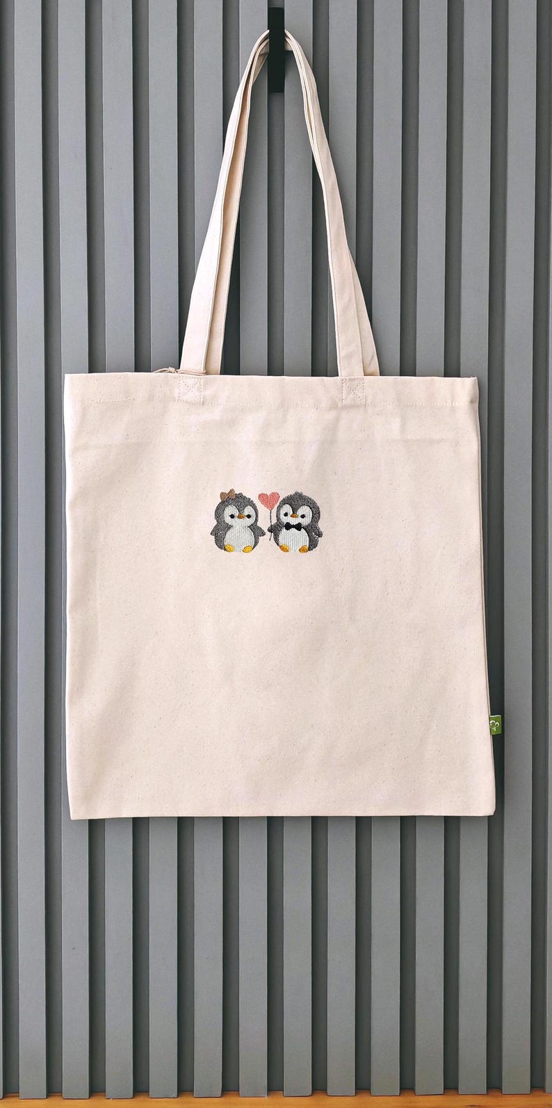 Puede incluir: Una bolsa de tela de color beige claro con un dise&ntilde;o bordado de dos peque&ntilde;os ping&uuml;inos de pie uno al lado del otro. Un ping&uuml;ino lleva un peque&ntilde;o lazo marr&oacute;n, el otro una pajarita negra, y sostienen un coraz&oacute;n rosa entre ambos.