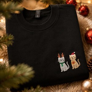 May include: A black crewneck sweatshirt featuring an embroidered design of two cats. One cat is light gray with brown reindeer antlers and a green scarf, while the other is tan with a red Santa hat and a small red heart on its chest.