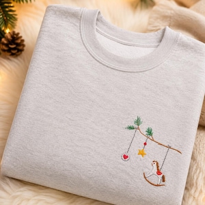 Puede incluir: Una sudadera gris claro de cuello redondo con un dise&ntilde;o bordado en el pecho. El bordado muestra una rama de &aacute;rbol marr&oacute;n con agujas de pino verdes, de la que cuelgan un adorno circular con un coraz&oacute;n rojo, una estrella amarilla y un caballito balanc&iacute;n.