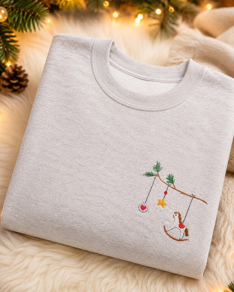 Puede incluir: Una sudadera gris claro de cuello redondo con un dise&ntilde;o bordado en el pecho. El bordado muestra una rama de &aacute;rbol marr&oacute;n con agujas de pino verdes, de la que cuelgan un adorno circular con un coraz&oacute;n rojo, una estrella amarilla y un caballito balanc&iacute;n.