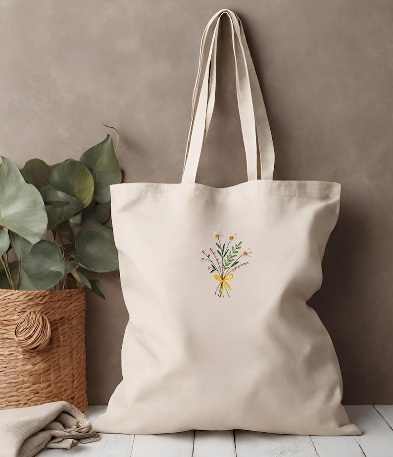 May include: A cream-colored canvas tote bag with a floral embroidery design. The embroidery features a bouquet of white flowers with green leaves and a yellow ribbon.
