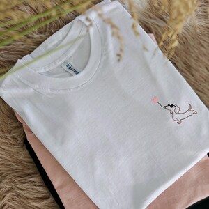 May include: White t-shirt with a pink embroidered dachshund holding a heart balloon.