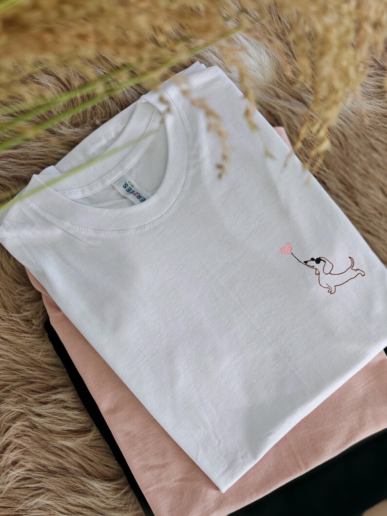 May include: White t-shirt with a pink embroidered dachshund holding a heart balloon.