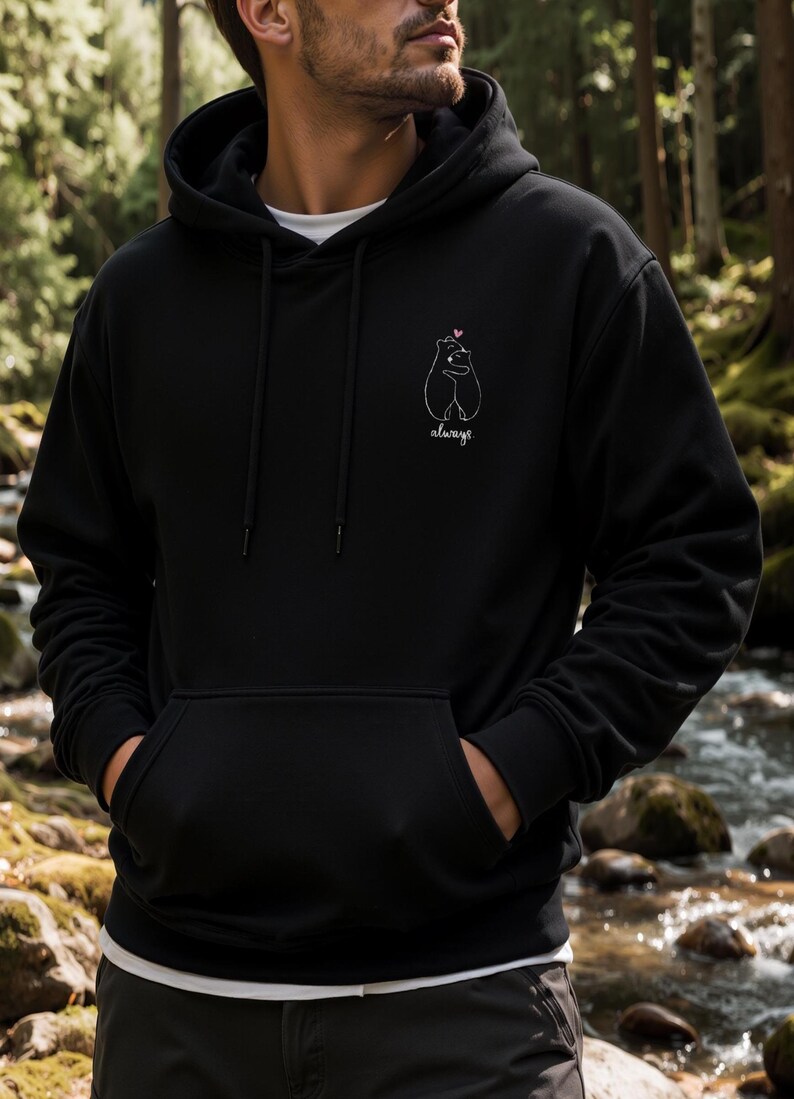 Op de afbeelding: Een zwarte hoodie met een witte lijntekening van twee beren die elkaar omhelzen met een klein roze hartje erboven en het woord always in cursief schrift onder de beren op de linkerzijde van de borst.