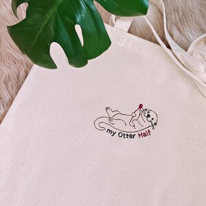 Puede incluir: Un bolso de tela color crema con un dise&ntilde;o bordado de una nutria marina tumbada boca arriba mientras sostiene un peque&ntilde;o coraz&oacute;n rojo. Debajo de la nutria, el texto "my Otter Half" est&aacute; bordado con hilo negro y rojo.
