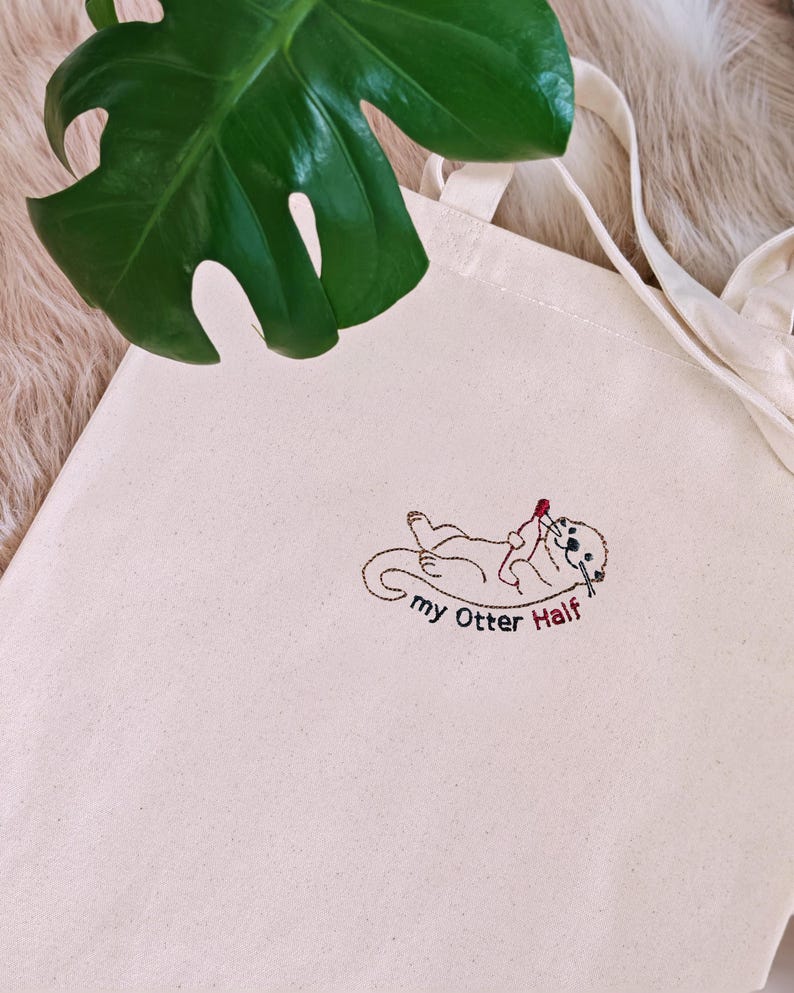 Puede incluir: Un bolso de tela color crema con un dise&ntilde;o bordado de una nutria marina tumbada boca arriba mientras sostiene un peque&ntilde;o coraz&oacute;n rojo. Debajo de la nutria, el texto "my Otter Half" est&aacute; bordado con hilo negro y rojo.