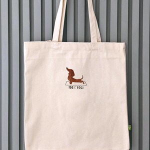 Puede incluir: Una bolsa de lona blanca con un perro salchicha marr&oacute;n bordado en ella haciendo una pose de yoga. El texto "100% YOGI" est&aacute; bordado debajo del perro.