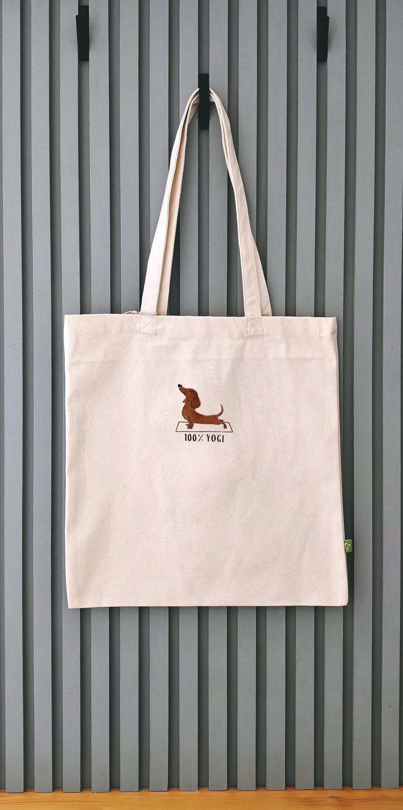 Puede incluir: Una bolsa de lona blanca con un perro salchicha marr&oacute;n bordado en ella haciendo una pose de yoga. El texto "100% YOGI" est&aacute; bordado debajo del perro.