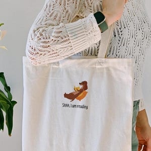 Puede incluir: Una bolsa de lona blanca con un dachshund marr&oacute;n bordado en ella. El dachshund est&aacute; acostado con un libro en sus patas. El texto "Shhh... I am reading" est&aacute; bordado debajo del dachshund.