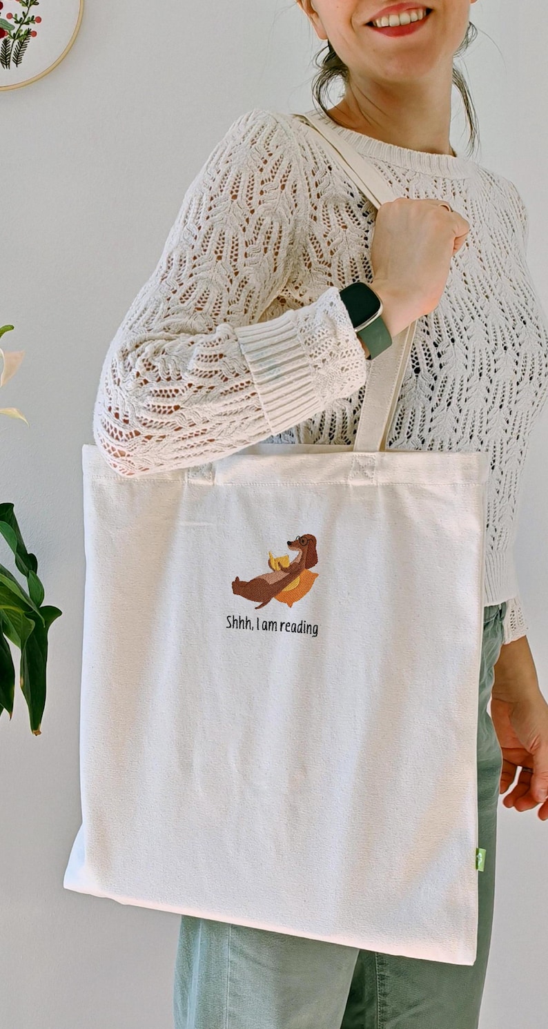 Puede incluir: Una bolsa de lona blanca con un dachshund marr&oacute;n bordado en ella. El dachshund est&aacute; acostado con un libro en sus patas. El texto "Shhh... I am reading" est&aacute; bordado debajo del dachshund.