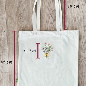 May include: A white canvas tote bag with a floral design and the letter "I" in red. The bag measures 38.5 inches wide, 42 inches tall, and 30 inches in height.