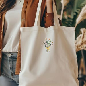 May include: White canvas tote bag with a small embroidered design of a bouquet of flowers tied with a yellow ribbon.