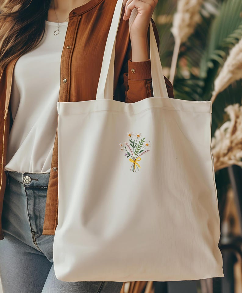 May include: White canvas tote bag with a small embroidered design of a bouquet of flowers tied with a yellow ribbon.