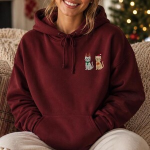 May include: A maroon hooded sweatshirt featuring an embroidered design on the chest of two cats. One cat wears brown reindeer antlers and a green scarf, while the other cat wears a red Santa hat and holds a small red ornament in its paw.