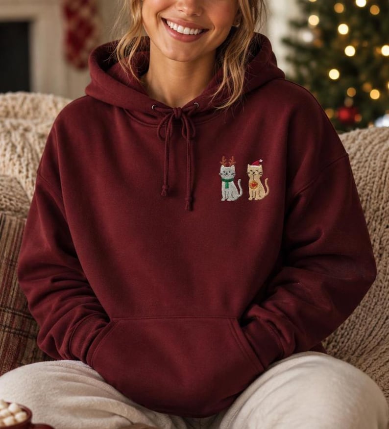 May include: A maroon hooded sweatshirt featuring an embroidered design on the chest of two cats. One cat wears brown reindeer antlers and a green scarf, while the other cat wears a red Santa hat and holds a small red ornament in its paw.