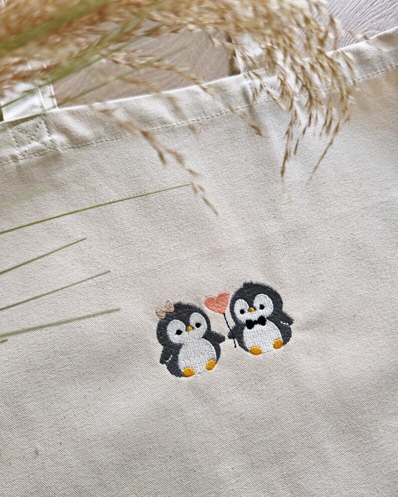 Puede incluir: Una bolsa de tela beige con un dise&ntilde;o bordado de dos peque&ntilde;os ping&uuml;inos. Un ping&uuml;ino lleva un peque&ntilde;o lazo, mientras que el otro lleva una pajarita. Est&aacute;n colocados uno al lado del otro, con uno sosteniendo un palo fino con un coraz&oacute;n rosa.