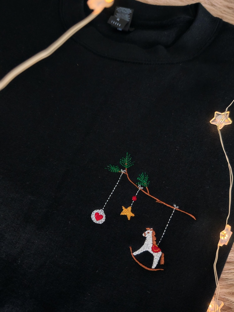 Könnte beinhalten: Schwarzes Sweatshirt mit Rundhalsausschnitt und gesticktem Design. Das Design zeigt einen Ast mit hängenden Ornamenten, darunter ein Herz, ein Stern und ein Schaukelpferd. Die Stickerei ist mehrfarbig.