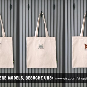 Puede incluir: Tres bolsas de tela de color beige claro con dos asas cada una. La bolsa de la izquierda muestra un perro salchicha y el texto Merry Woofmas. La bolsa central muestra una cara de gato y el texto Fluff You. La derecha muestra un perro salchicha y el texto 100% YOU.