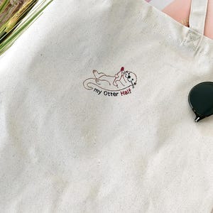 Puede incluir: Un bolso de tela color crema con un bordado que muestra una nutria tumbada boca arriba sosteniendo un coraz&oacute;n rojo. Debajo de la nutria, el texto "my Otter Half" est&aacute; bordado en hilo marr&oacute;n oscuro. El bolso tiene dos asas largas.