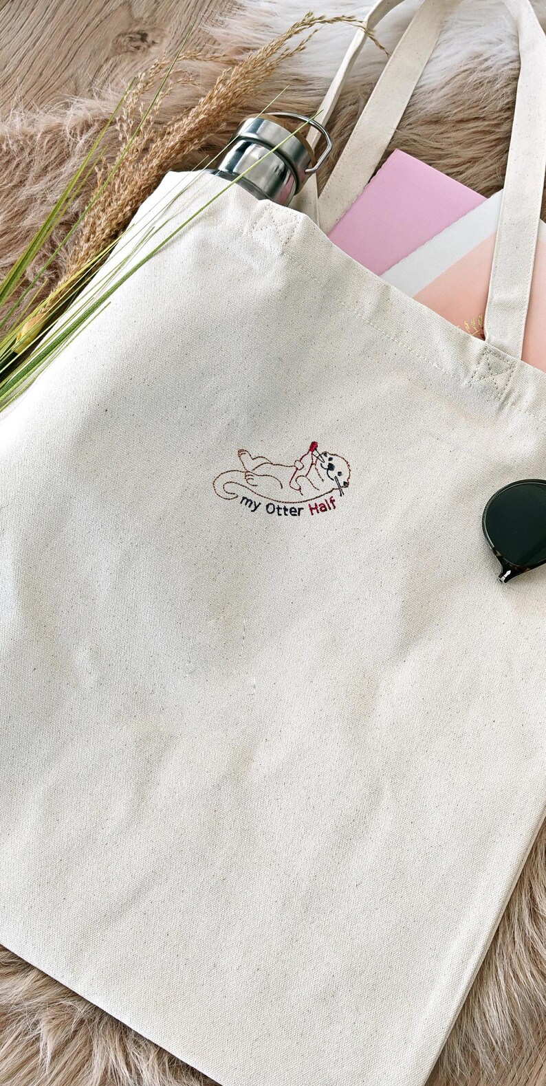 Puede incluir: Un bolso de tela color crema con un bordado que muestra una nutria tumbada boca arriba sosteniendo un coraz&oacute;n rojo. Debajo de la nutria, el texto "my Otter Half" est&aacute; bordado en hilo marr&oacute;n oscuro. El bolso tiene dos asas largas.