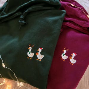 May include: Two hooded sweatshirts, one dark green and one burgundy, are displayed. Each sweatshirt features an embroidered design of two geese wearing festive holiday accessories. The geese are adorned with reindeer antlers, scarves, and Santa hats.