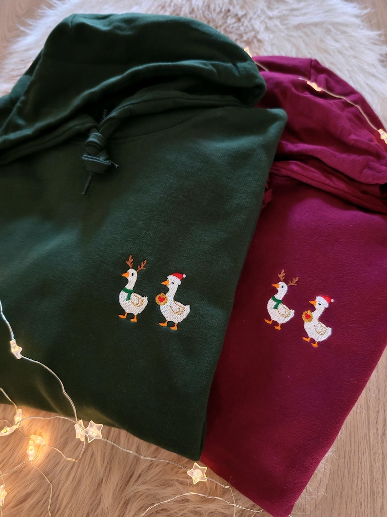 May include: Two hooded sweatshirts, one dark green and one burgundy, are displayed. Each sweatshirt features an embroidered design of two geese wearing festive holiday accessories. The geese are adorned with reindeer antlers, scarves, and Santa hats.