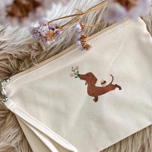 May include: Cream-colored canvas clutch with a brown dachshund embroidery carrying flowers. The clutch has a wrist strap and a zipper closure. The background is a fluffy, light-colored surface.