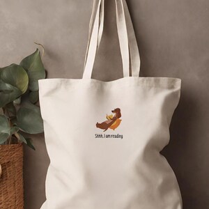 Puede incluir: Una bolsa de lona blanca con un perro salchicha marr&oacute;n leyendo un libro. El texto en la bolsa dice "Shhh, estoy leyendo."