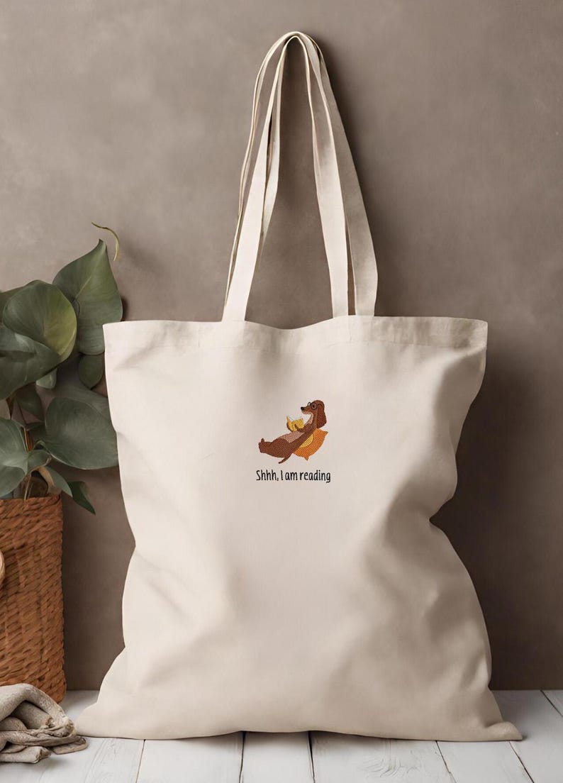 Puede incluir: Una bolsa de lona blanca con un perro salchicha marr&oacute;n leyendo un libro. El texto en la bolsa dice "Shhh, estoy leyendo."