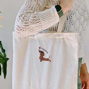 May include: Cream-colored tote bag with a dachshund illustration holding a wine glass and the word "Cheers!". The bag has long handles and is made of a sturdy fabric, suitable for carrying groceries or other items.