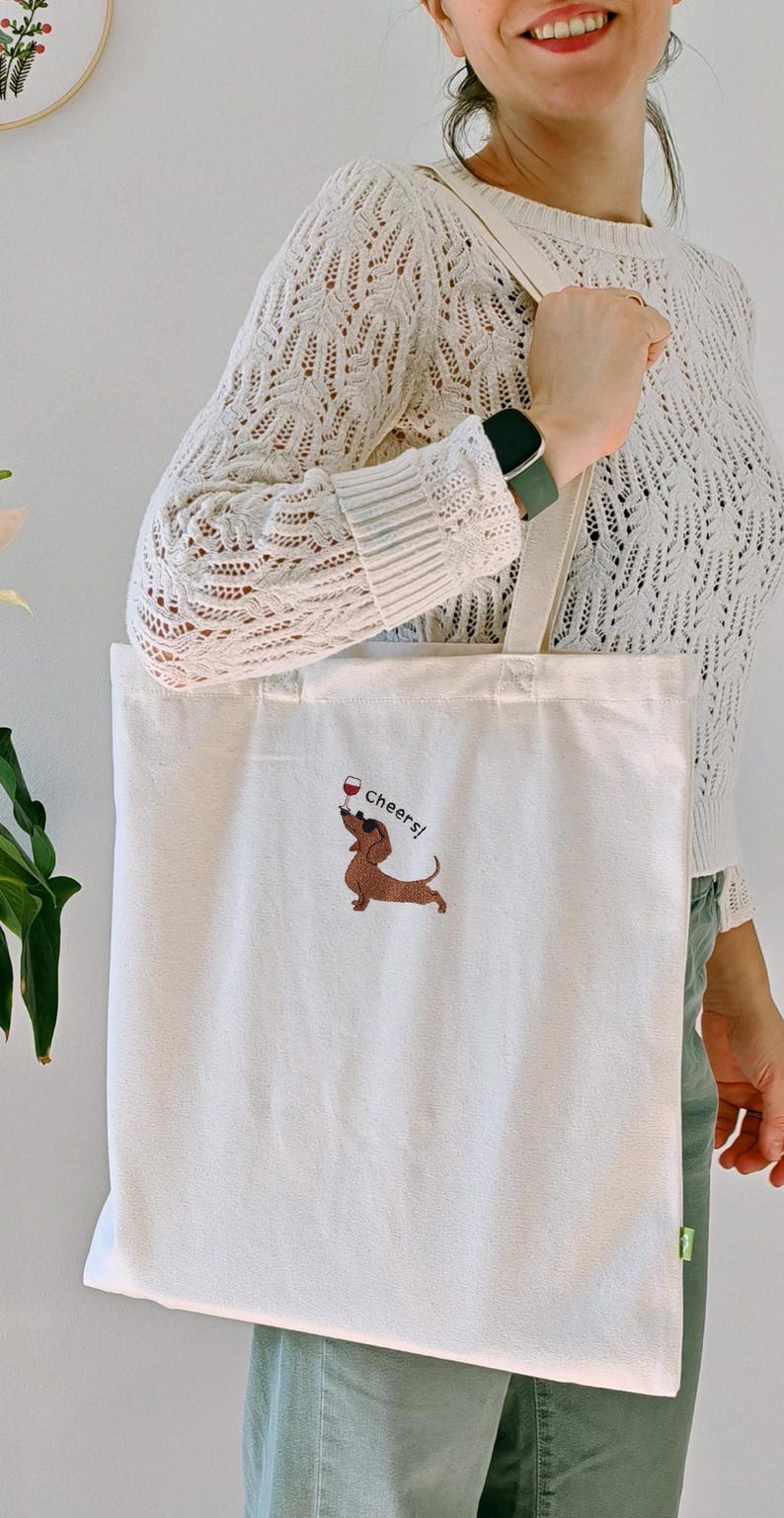 May include: Cream-colored tote bag with a dachshund illustration holding a wine glass and the word "Cheers!". The bag has long handles and is made of a sturdy fabric, suitable for carrying groceries or other items.