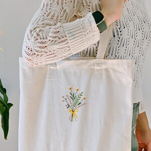 May include: A white canvas tote bag with a small embroidered bouquet of yellow and green flowers. The flowers are tied with a yellow ribbon.