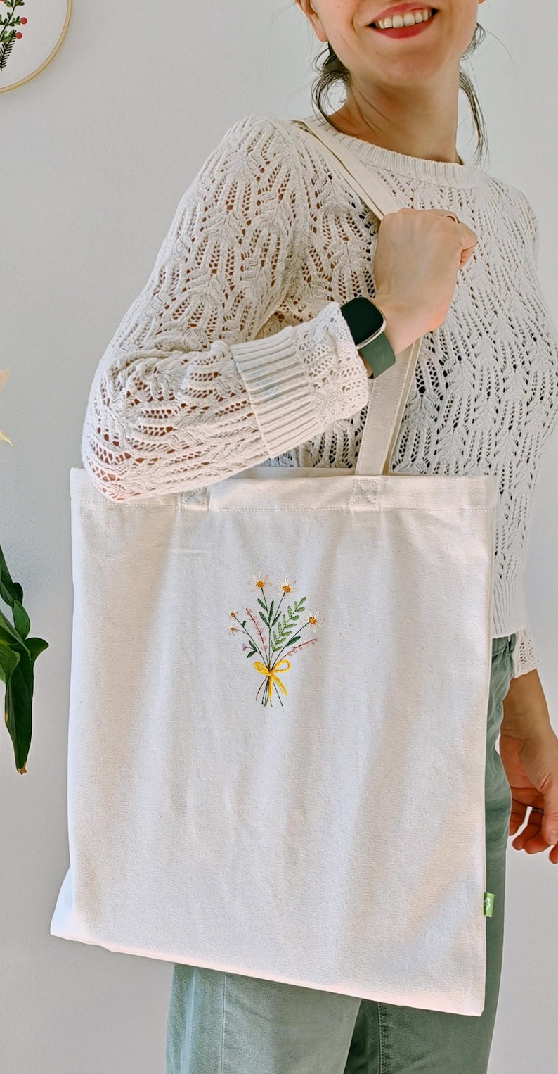 May include: A white canvas tote bag with a small embroidered bouquet of yellow and green flowers. The flowers are tied with a yellow ribbon.