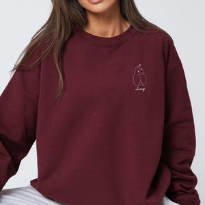 Op de afbeelding: Een diepbordeauxrood sweatshirt met lange mouwen en een ronde hals, voorzien van een kleine witte lijntekening op de linkerborst. De afbeelding toont twee beren die elkaar omhelzen met een klein hartje erboven en het woord always in cursief.