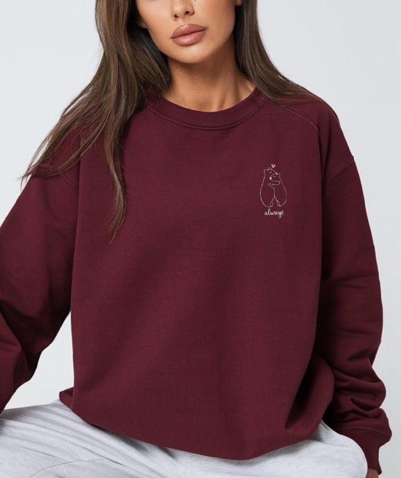 Op de afbeelding: Een diepbordeauxrood sweatshirt met lange mouwen en een ronde hals, voorzien van een kleine witte lijntekening op de linkerborst. De afbeelding toont twee beren die elkaar omhelzen met een klein hartje erboven en het woord always in cursief.