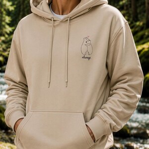 Op de afbeelding: Een beige hoodie met een kangoeroezak aan de voorzijde, lange mouwen en een kleine zwarte lijntekening op de borst van twee knuffelende beren met een klein roze hartje erboven en het woord always in een cursief handschrift geschreven.