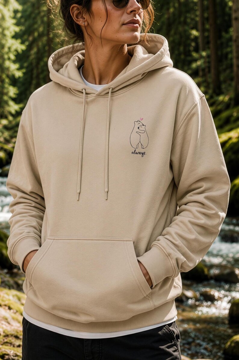 Op de afbeelding: Een beige hoodie met een kangoeroezak aan de voorzijde, lange mouwen en een kleine zwarte lijntekening op de borst van twee knuffelende beren met een klein roze hartje erboven en het woord always in een cursief handschrift geschreven.