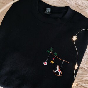 Puede incluir: Una sudadera negra de cuello redondo con un dise&ntilde;o bordado en el pecho. El bordado muestra una rama marr&oacute;n con agujas de pino verdes, de la que cuelgan un adorno rojo y blanco, una estrella amarilla y un caballo balanc&iacute;n blanco.
