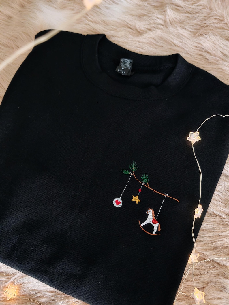 Puede incluir: Una sudadera negra de cuello redondo con un dise&ntilde;o bordado en el pecho. El bordado muestra una rama marr&oacute;n con agujas de pino verdes, de la que cuelgan un adorno rojo y blanco, una estrella amarilla y un caballo balanc&iacute;n blanco.