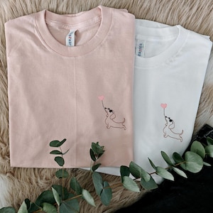 May include: Two t-shirts, one pink and one white, with an embroidered image of a dachshund holding a heart-shaped balloon. The shirts have the brand name "ERZEES" printed on the tag.