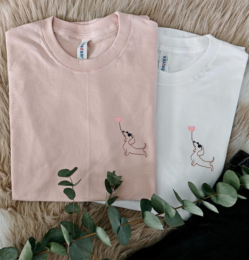 May include: Two t-shirts, one pink and one white, with an embroidered image of a dachshund holding a heart-shaped balloon. The shirts have the brand name "ERZEES" printed on the tag.