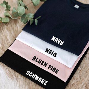 May include: Four t-shirts stacked on top of each other. The top shirt is navy blue, the next is white, the next is blush pink, and the bottom shirt is black. The shirts have the words "NAVY", "WEISS", "BLUSH PINK", and "SCHWARZ" printed on them.