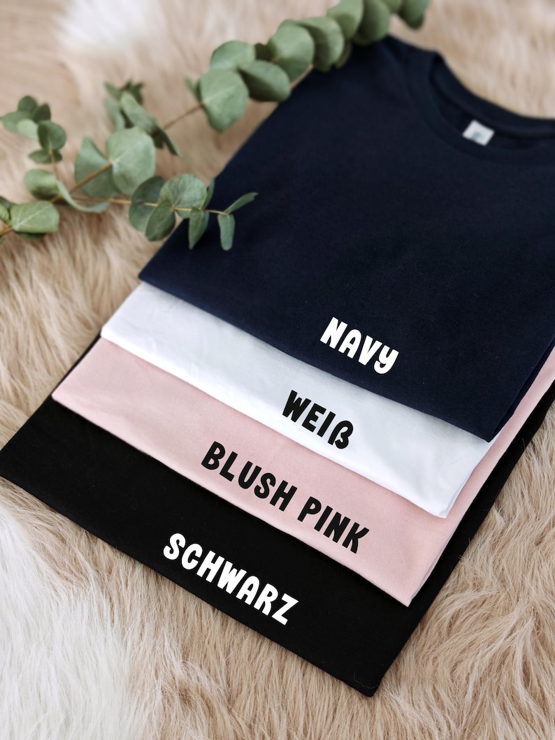 May include: Four t-shirts stacked on top of each other. The top shirt is navy blue, the next is white, the next is blush pink, and the bottom shirt is black. The shirts have the words "NAVY", "WEISS", "BLUSH PINK", and "SCHWARZ" printed on them.