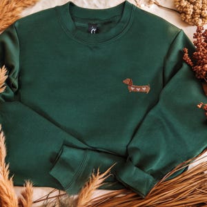 Süßer Dackel Pullover als Geschenk für Dackelliebhaber
Bestickter Hunde Hoodie für Hundemama und Hundepapa Ein dunkelgrünes Sweatshirt mit Rundhalsausschnitt und langen Ärmeln. Auf der linken Brust befindet sich ein kleiner gestickter Dackel mit Herzakzenten. Das Sweatshirt ist aus einem weichen Material.