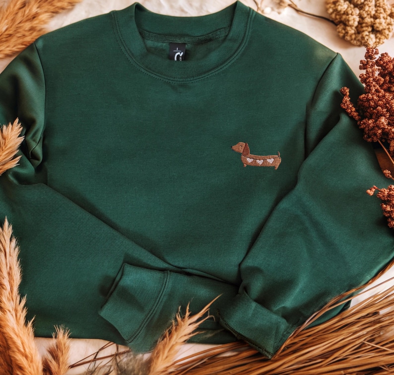 Süßer Dackel Pullover als Geschenk für Dackelliebhaber
Bestickter Hunde Hoodie für Hundemama und Hundepapa Ein dunkelgrünes Sweatshirt mit Rundhalsausschnitt und langen Ärmeln. Auf der linken Brust befindet sich ein kleiner gestickter Dackel mit Herzakzenten. Das Sweatshirt ist aus einem weichen Material.