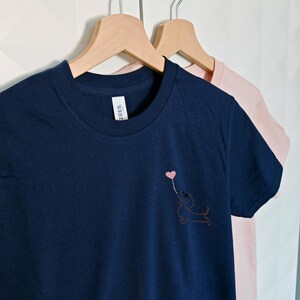 May include: Two t-shirts hanging on wooden hangers. The shirt on the left is navy blue with a small embroidered design of a dachshund holding a heart-shaped balloon. The shirt on the right is pink.
