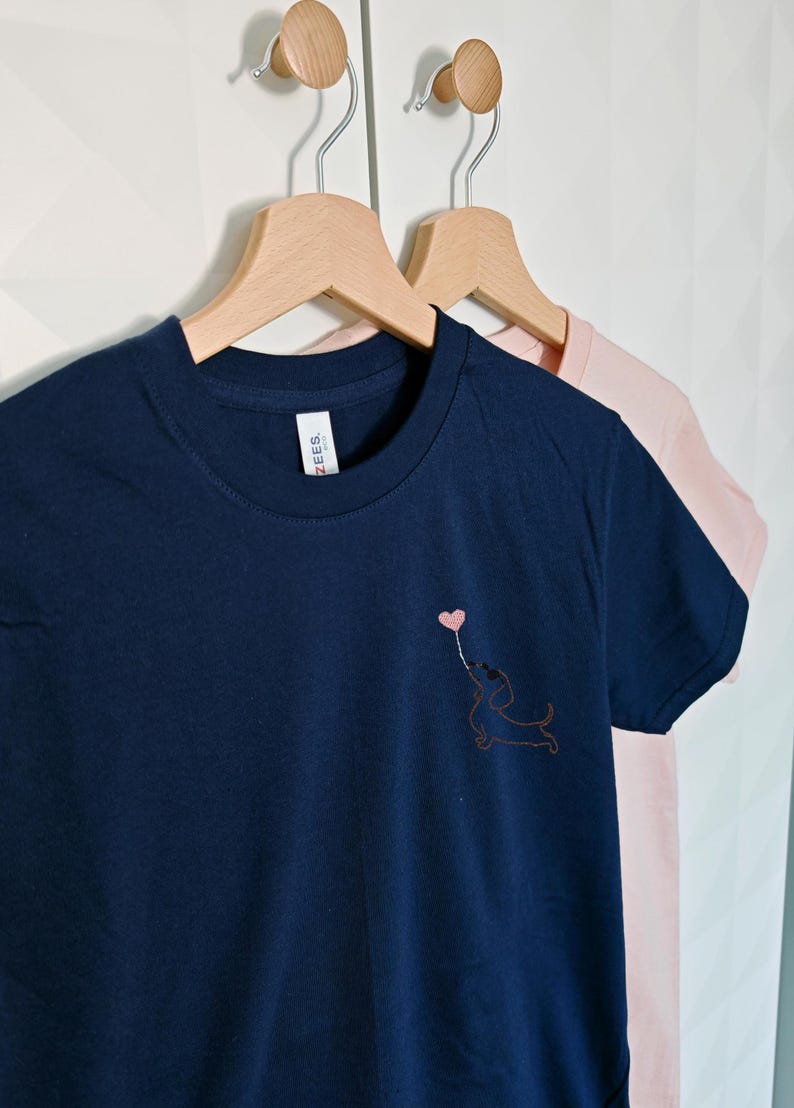 May include: Two t-shirts hanging on wooden hangers. The shirt on the left is navy blue with a small embroidered design of a dachshund holding a heart-shaped balloon. The shirt on the right is pink.