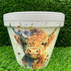 May include: A white ceramic plant pot decorated with a watercolour image of a Highland cow wearing a floral crown. The pot has a textured rim and a tapered shape. The cow's fur is brown and the flowers are purple and blue.