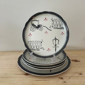 May include: Set of four white ceramic plates with black and red designs. The plates feature a black carriage, a street lamp, and a fence.