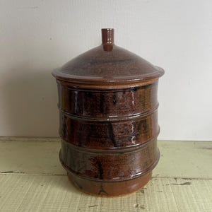 May include: A brown ceramic jar with a lid. The jar has a textured surface and is decorated with black lines. The lid is slightly domed and has a small knob on top.